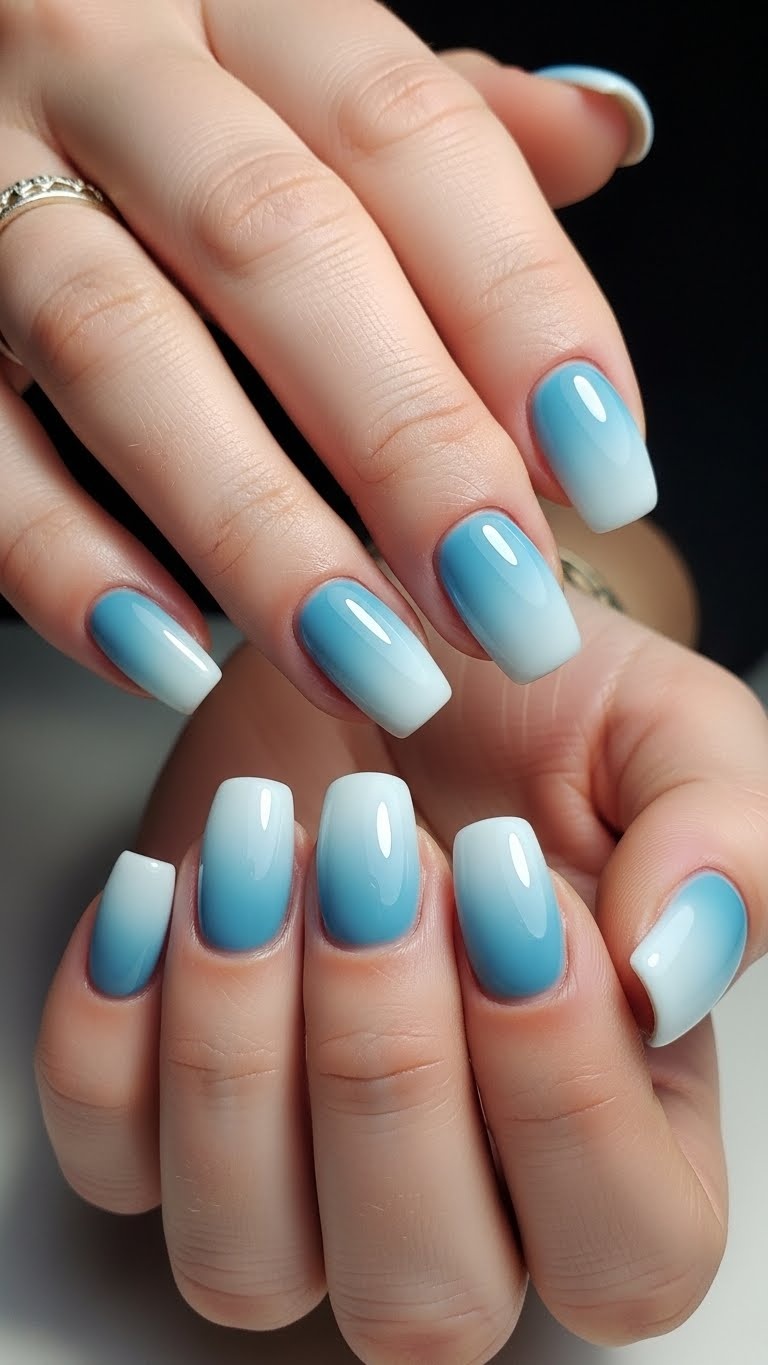 Close-up of hands with nails painted in a smooth icy blue ombre gradient, posed against a soft winter-themed background.