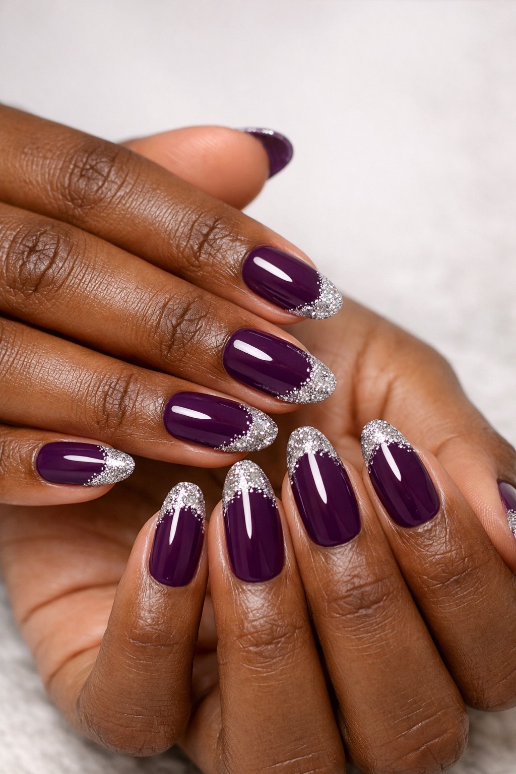 Close-up of hands with dark skin tones showing nails painted plum purple with silver glitter tips.