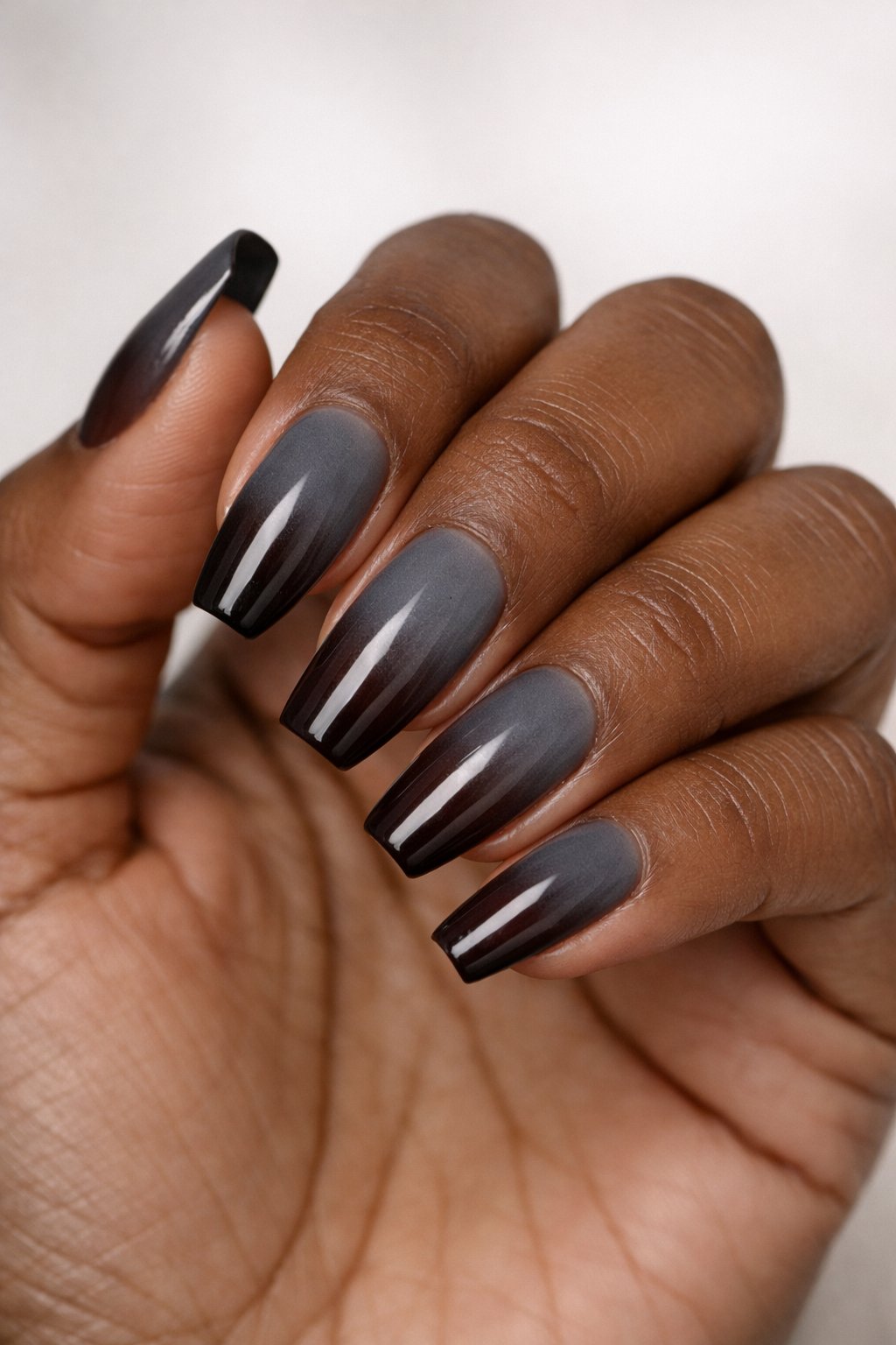Close-up of a hand with dark skin showing nails painted in smoky gray ombre fading into deep brown.