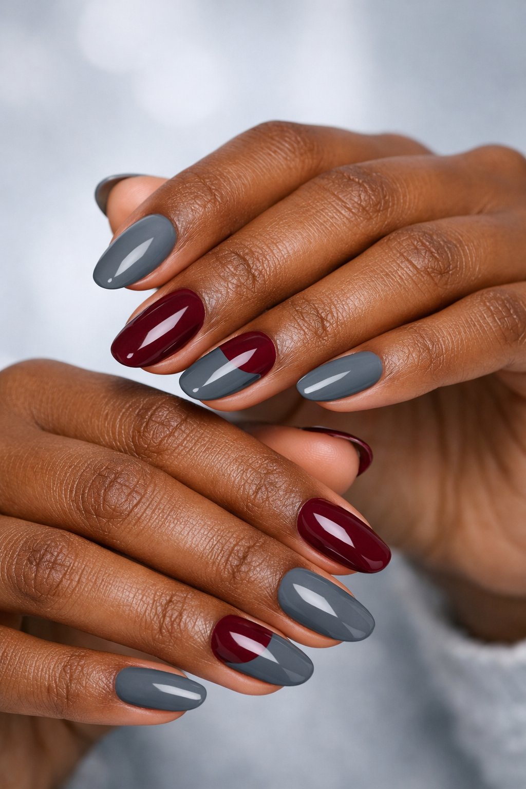 Close-up of dark-skinned hands with gray and burgundy painted nails posed against a neutral background.