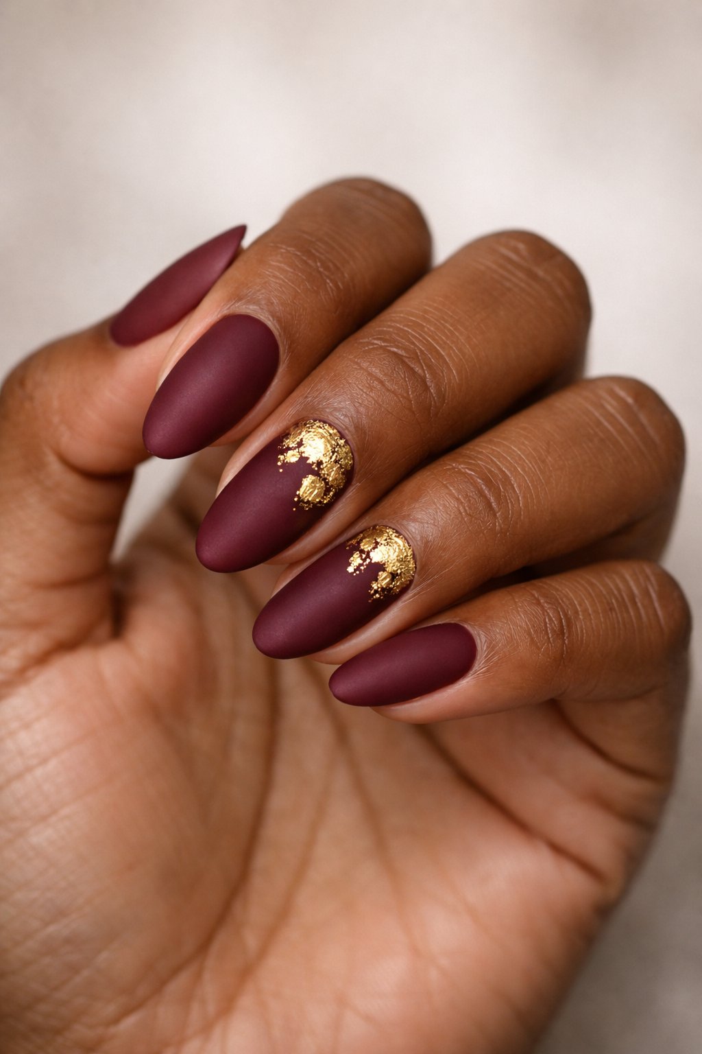 Close-up of a dark-skinned hand with deep burgundy matte nails and gold foil accents.