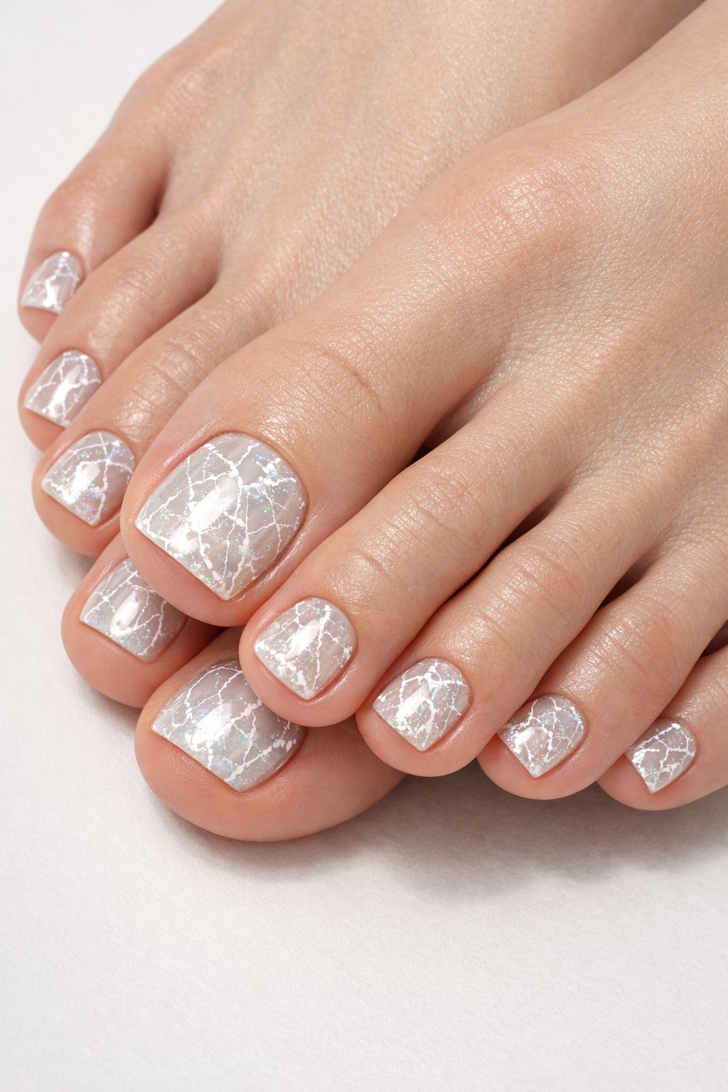 Close-up of a woman's feet with white and clear cracked ice effect toenail polish on a neutral background.