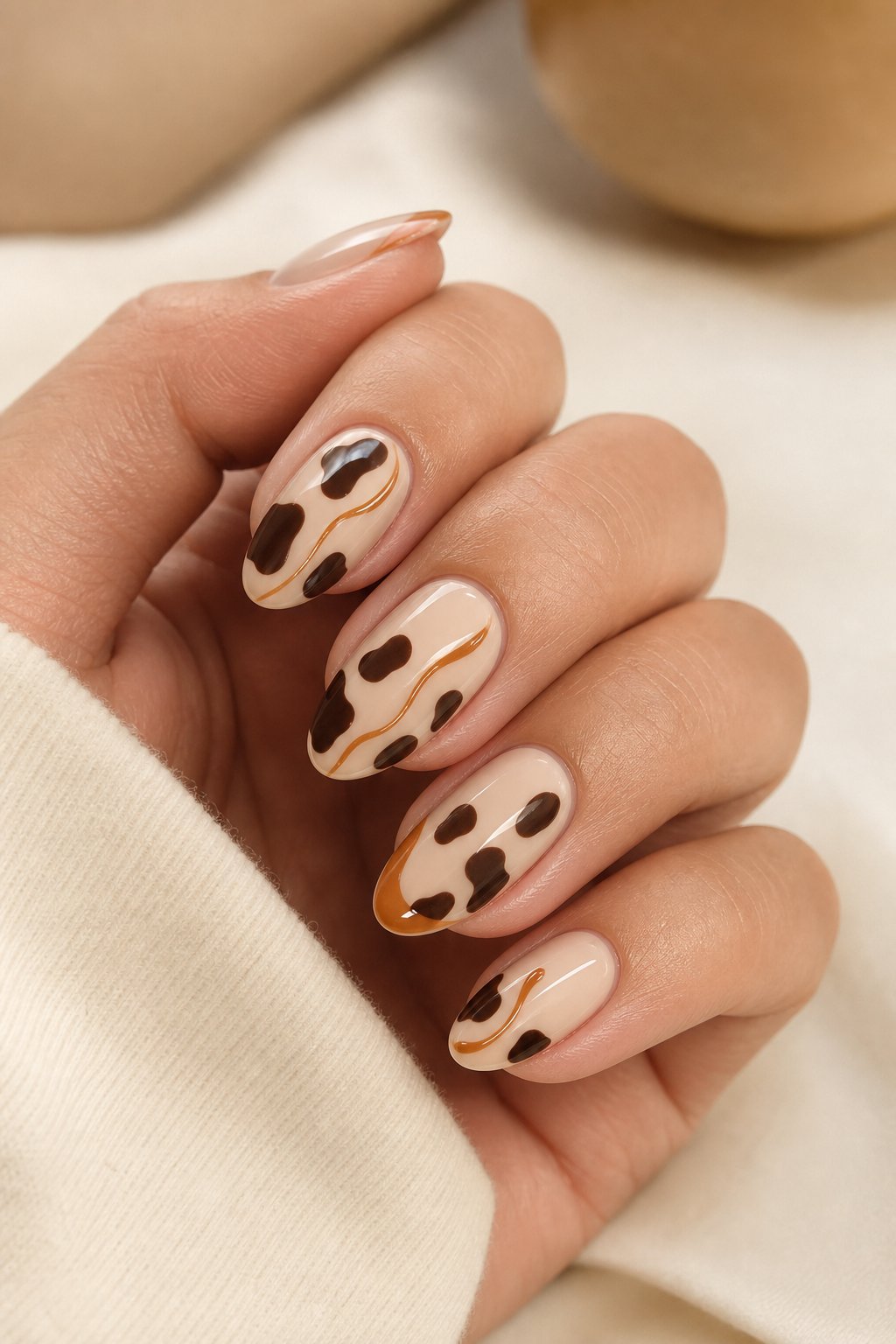A hand with glossy almond-shaped nails featuring brown cow-print patterns and caramel accents.